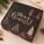 Czarne pudełko świąteczne ze złotym napisem Merry Christmas i dekoracyjnymi choinkami.